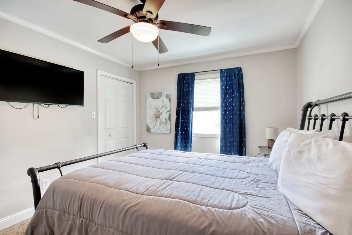 A warmly decorated bedroom with a plush bed, soft curtains framing the window, elegant wall art, and a television. The natural light streaming in sets a tranquil mood, perfect for unwinding or enjoying a quiet night in.
