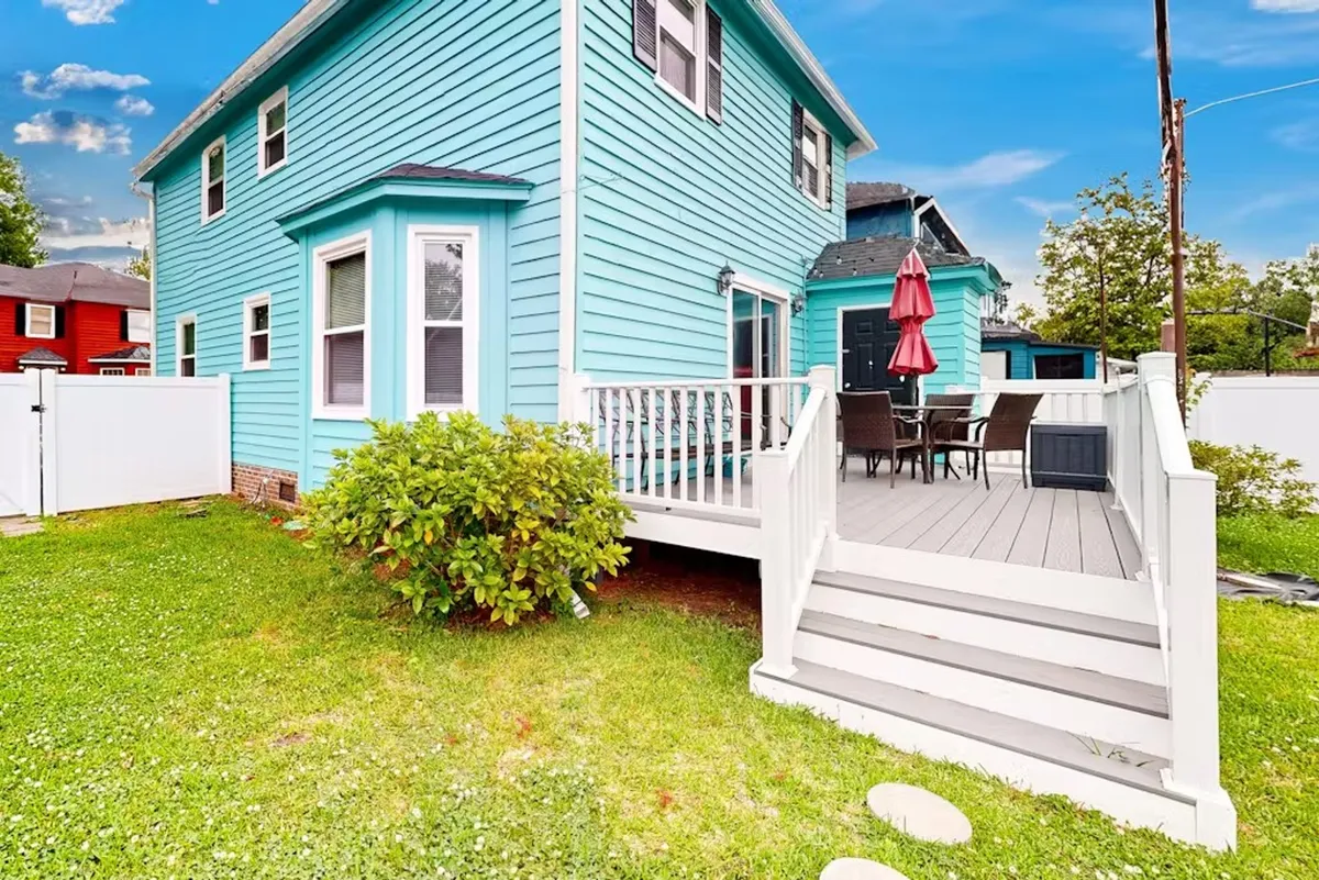 An inviting deck with a bright blue house in the background, white railing, red patio umbrella, and a dining set arranged for outdoor meals. The deck overlooks a spacious, sunny lawn—perfect for play or relaxation.
