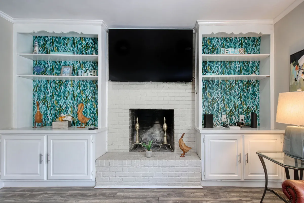 A stylish living area with a television above a modern fireplace, decorative shelves, and vibrant, colorful wallpaper. The space is arranged for both relaxation and group gatherings, ideal for a warm, social atmosphere.