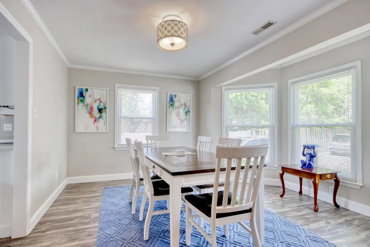 A welcoming dining area centered around a large table, surrounded by artwork and illuminated by a distinctive light fixture. Sunlight streams in from nearby windows, creating a bright and lively setting ideal for family dinners or special occasions.