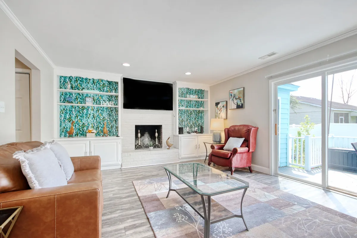 A cozy living room scene featuring a striking fireplace, green wallpaper adorned with lush plant motifs, a vibrant red armchair, and a glass coffee table. The space is warmly lit, inviting guests to gather and enjoy conversation or plan an evening out.