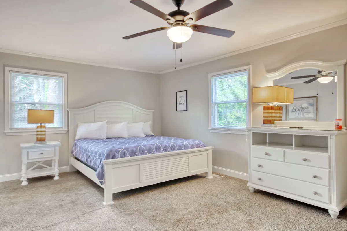 A cozy bedroom featuring a comfortable bed, dresser, lamps, and sunlit windows. The ambiance is relaxed, inviting you to unwind or enjoy a leisurely morning in bed.