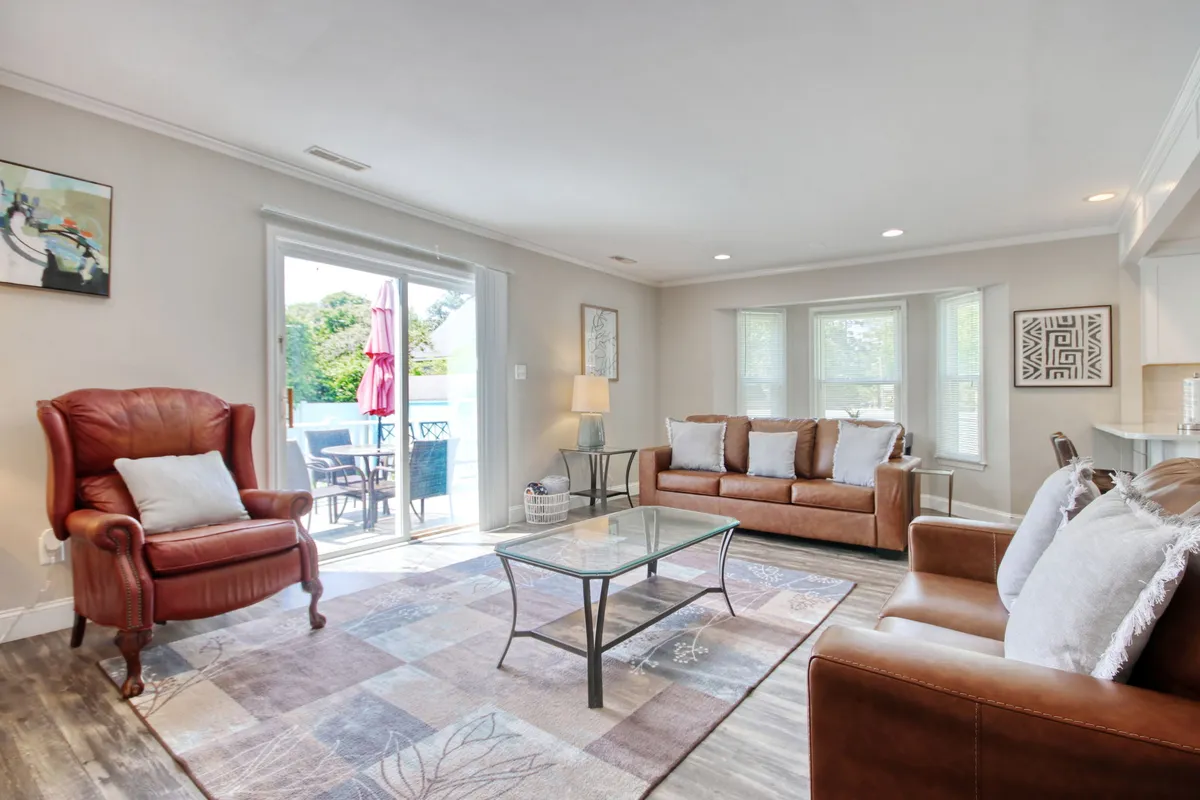 A bright living room anchored by a red armchair and a comfortable sofa with cushions arranged around a glass coffee table. Large windows flood the space with natural light, highlighting the tasteful decor and cheerful atmosphere. The setup invites relaxation, conversation, and casual gatherings.