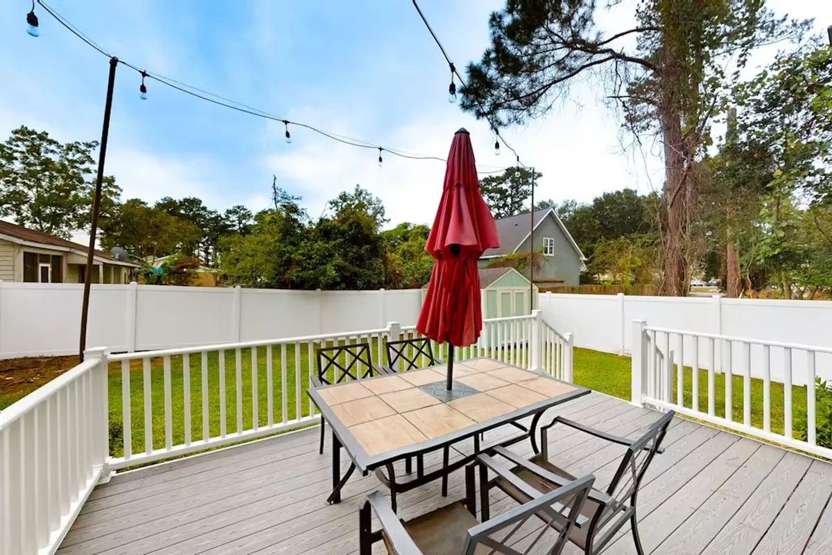 A backyard setting with a large outdoor dining table under an umbrella, string lights overhead, a green lawn, and a fenced yard. The space is set for family gatherings, social meals, and play, with children visible on the grass.