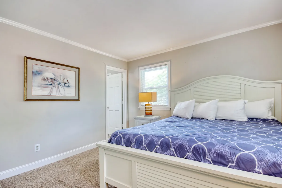 A serene bedroom with a plush bed, soft linens, a calming painting above the headboard, a bedside lamp casting gentle light, and a window that lets in natural light. The space is uncluttered and thoughtfully decorated, emphasizing tranquility and relaxation.