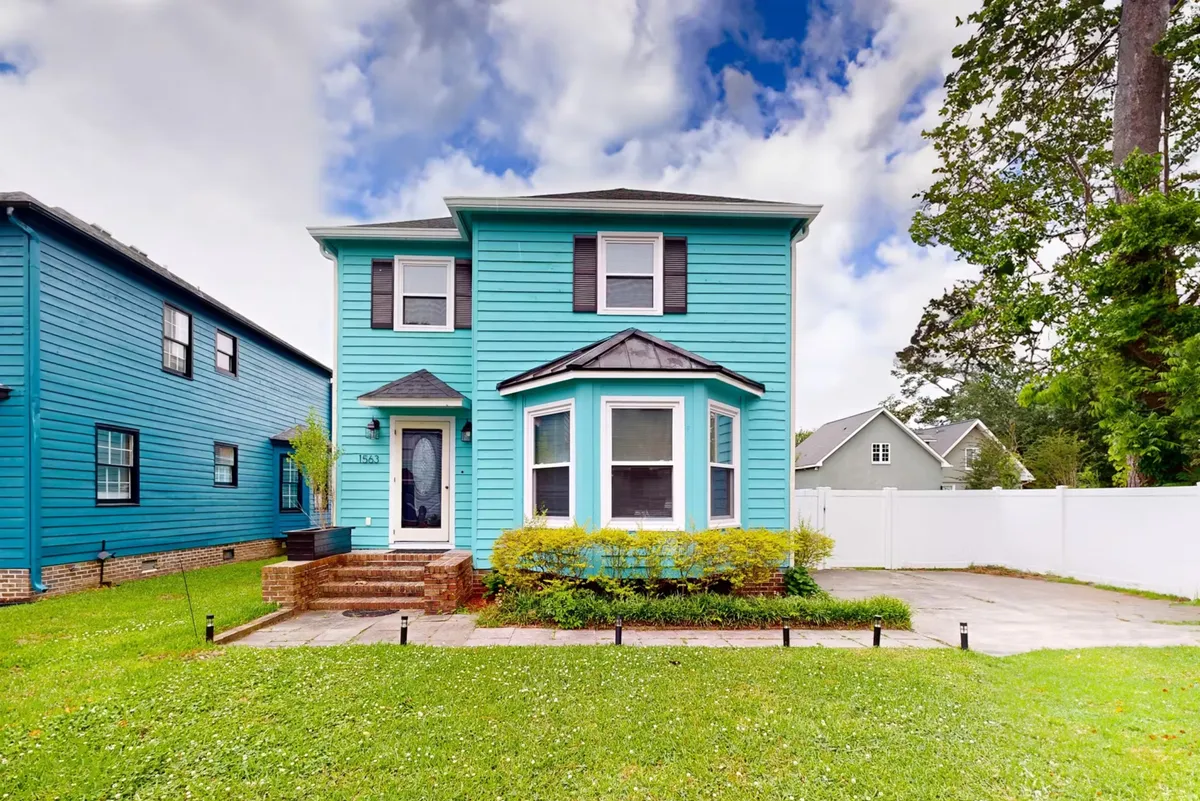 The image showcases the bright turquoise exterior of a single-family house in Myrtle Beach, with large front windows, a meticulously kept lawn, and a welcoming front porch perfect for sunset views or morning coffee. The inviting atmosphere suggests space for family gatherings and outdoor relaxation.