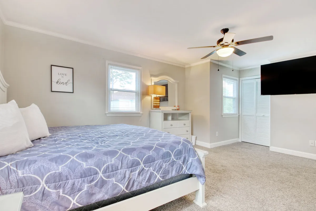 A tranquil bedroom scene featuring a comfortable bed with plush linens, a nightstand with a soft lamp, a dresser, and wall art that adds a touch of personality. Natural light filters in, creating a peaceful atmosphere ideal for relaxing mornings or intimate evenings. A TV on the dresser offers entertainment options, and the overall room feels cozy and inviting.